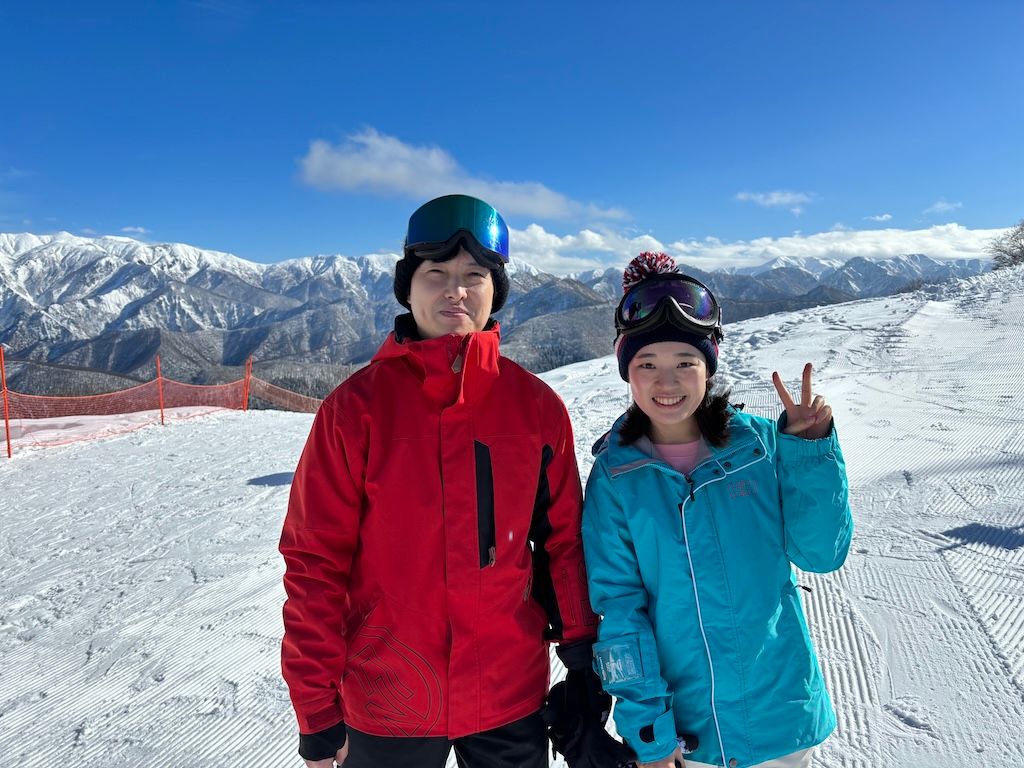 Arisa and Yuichiro enjoying a ski trip together on a snowy mountain slope with beautiful alpine scenery in the background