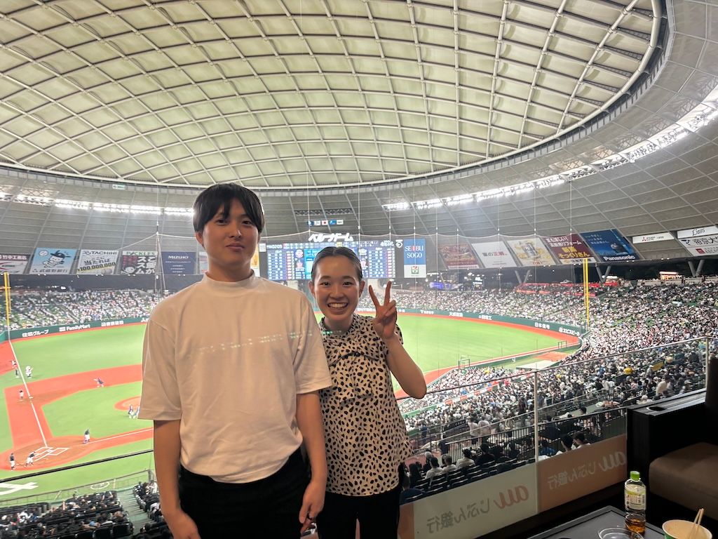 Arisa and Yuichiro at a baseball stadium, smiling and making peace signs while enjoying a game together