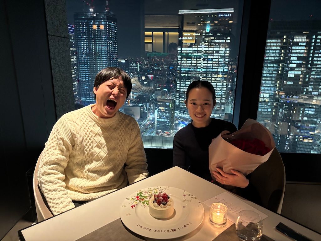 Arisa and Yuichiro celebrating at a romantic restaurant with city night view, sharing a special moment with a dessert and flowers