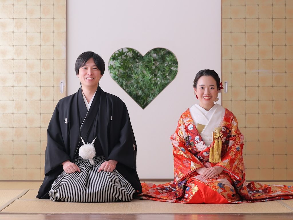Traditional Japanese wedding portrait of Arisa in vibrant red uchikake kimono and Yuichiro in formal black montsuki, sitting in traditional tatami room with heart-shaped greenery backdrop